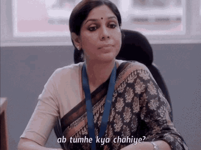 a woman sitting at a desk with the words ab tumhe kya chahiye written below her