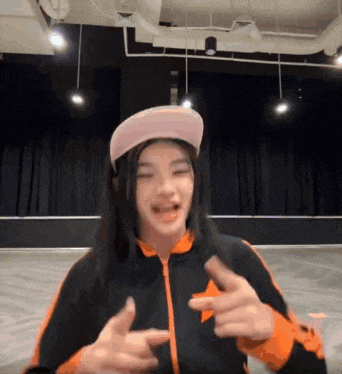 a girl wearing a pink hat and an orange jacket is pointing at the camera