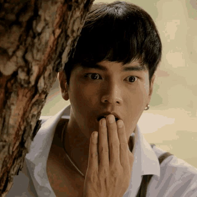 a young man covering his mouth with his hand while standing next to a tree