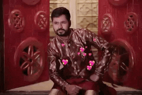 a man with a beard is sitting in front of a red wall with hearts coming out of his shirt .