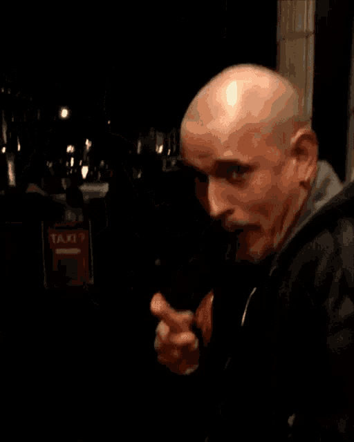 a bald man in a black jacket is pointing at the camera