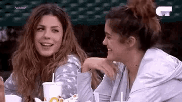 two women are sitting at a table talking to each other while drinking soda .