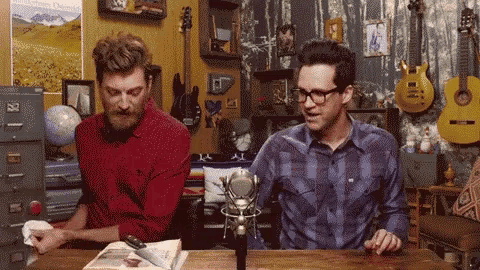 two men are sitting at a table in front of a microphone in a room with guitars .