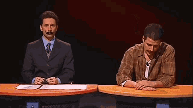 a man in a suit and tie sits at a table next to another man in a plaid shirt
