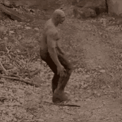 a man without a shirt is standing in the dirt holding a hammer .
