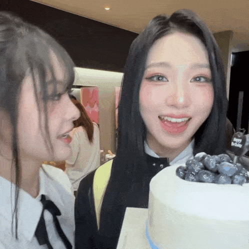 two girls standing next to a cake with blueberries on top