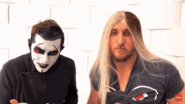 a man with long blonde hair is standing next to a man with a black shirt that says subscribe