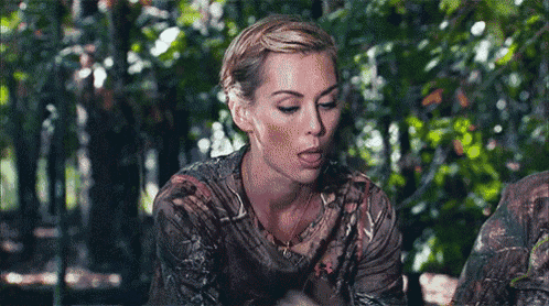 a woman in a camo shirt is sitting in the woods eating a piece of food .