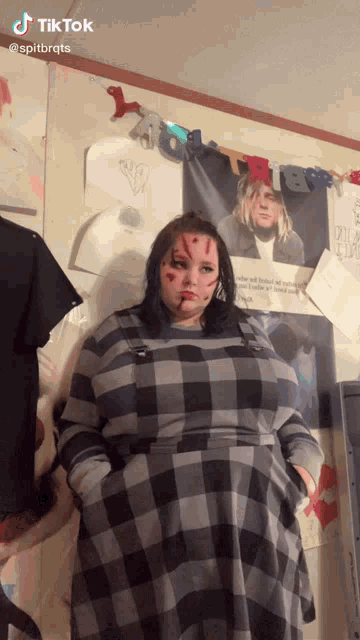 a woman in a plaid dress is standing in front of a wall with a picture of kurt cobain hanging on it