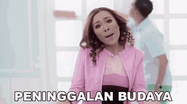a woman in a pink jacket is standing in front of a man in a blue shirt and a sign that says peninggalan budaya