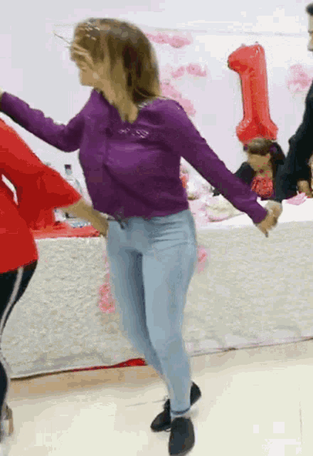 a woman in a purple shirt and blue jeans is dancing in front of a number 1 balloon