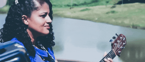 a woman with curly hair is holding a guitar in front of a lake