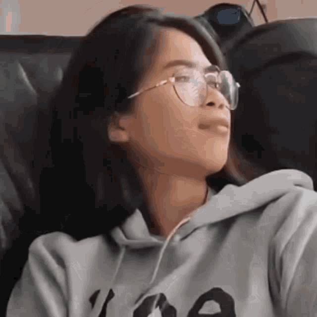 a girl wearing glasses and a hoodie is sitting on a couch .