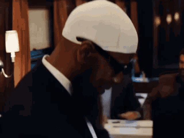 a man wearing a white hat and sunglasses is sitting at a table in a dark room .