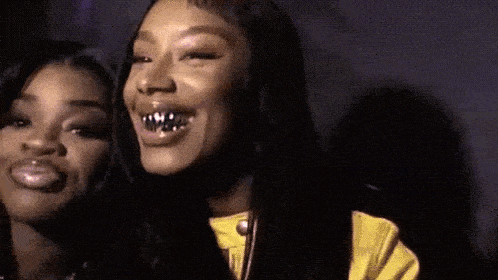 two women are standing next to each other and smiling . one of the women has a grill on her teeth .
