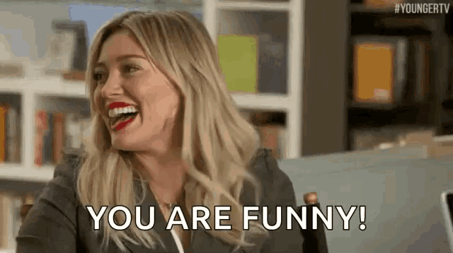 a woman is laughing and saying `` you are funny '' while sitting in a chair in front of a bookshelf .