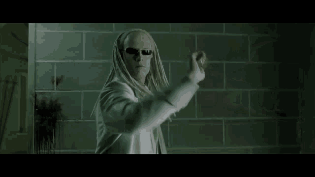 a man with dreadlocks and sunglasses is standing in front of a brick wall in a room .
