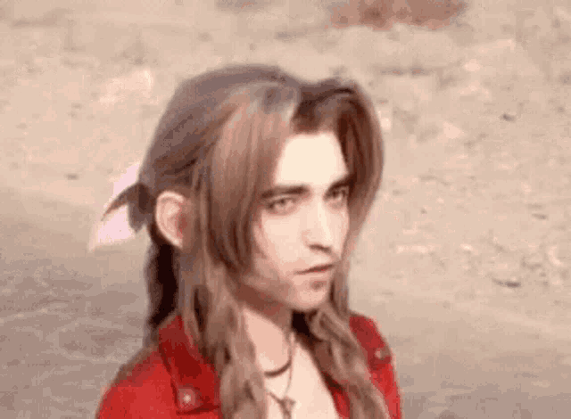 a man with long hair and a red shirt is standing in the sand .