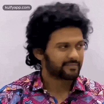 a man with curly hair and a beard is wearing a colorful shirt and looking at the camera .