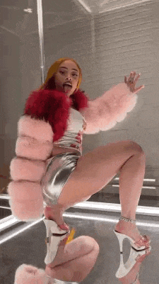 a woman in a pink fur coat and silver shorts is kneeling down in front of a mirror and sticking her tongue out .