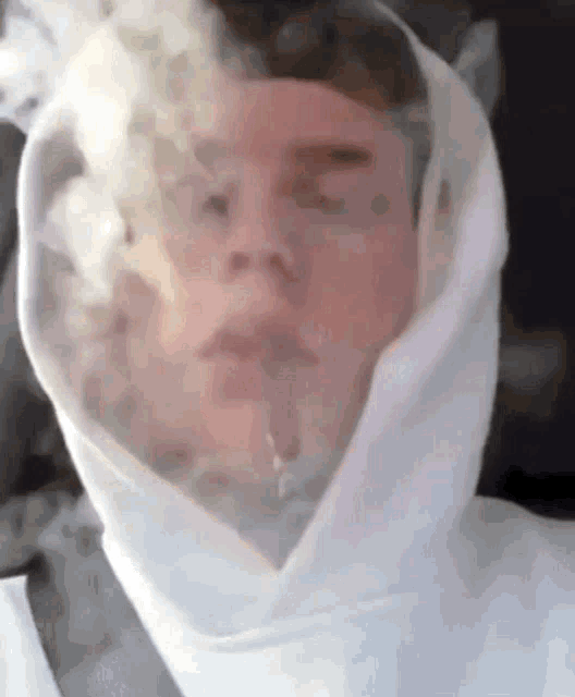 a man in a white hoodie is smoking a cigarette with smoke coming out of his face .