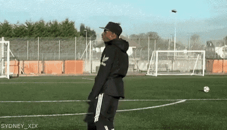 a man in a black adidas jacket is standing on a soccer field looking at a drone flying overhead .