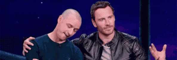 two men are sitting next to each other on a stage . one of the men has a bald head .