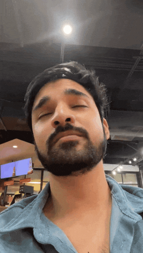 a man with a beard wearing a blue shirt is looking at the camera with his eyes closed