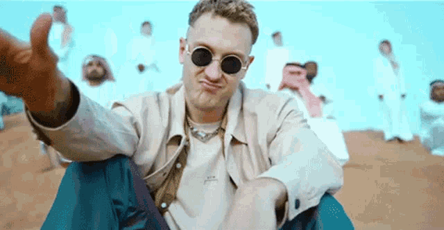 a man wearing sunglasses is sitting on the ground in front of a crowd of people .