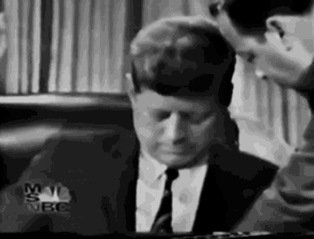 a black and white photo of a man in a suit and tie sitting at a table .