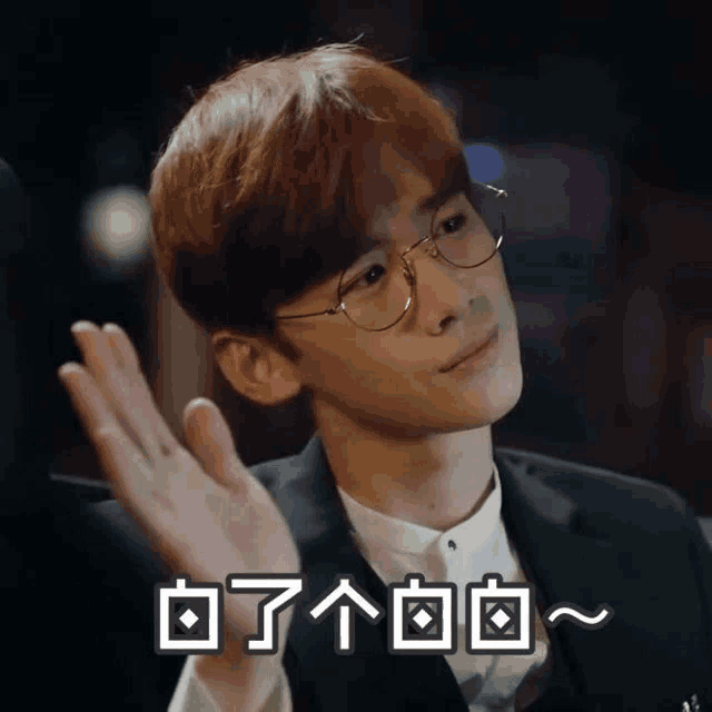 a young man wearing glasses and a suit is waving his hand in a foreign language .