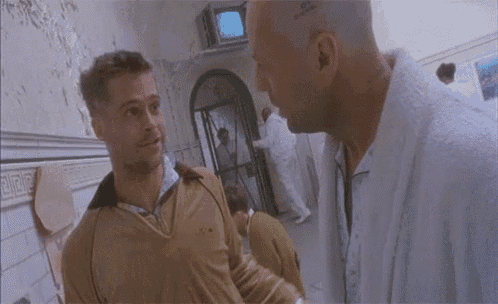two men are standing next to each other in a hallway and talking . one of the men has a tattoo on his head .
