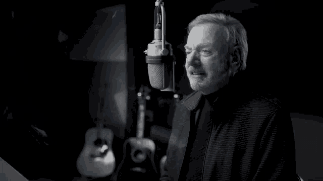 a man singing into a microphone with a guitar behind him