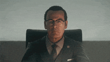 a man in a suit and tie sits in a chair with a badge on his chest
