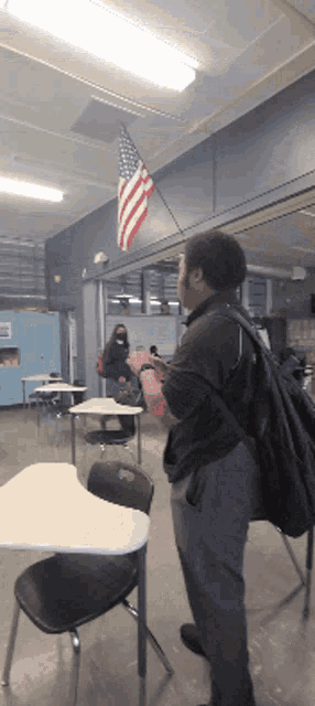 a man with a backpack stands in a classroom with an american flag hanging from the ceiling