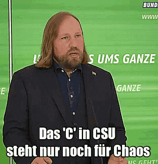 a man with long hair and a beard stands in front of a green background that says ' ganze ' on it