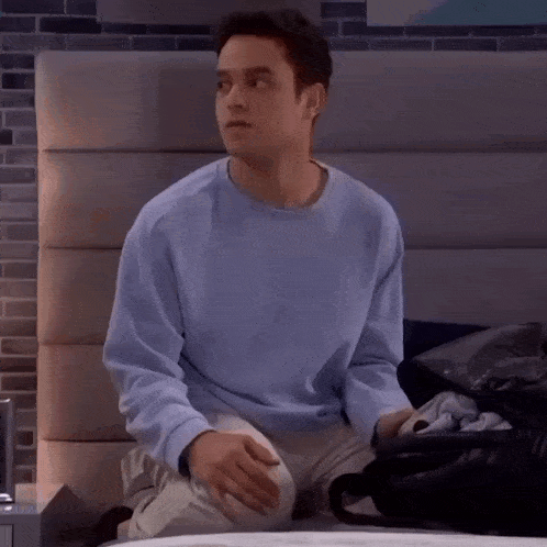 a man in a blue sweater is sitting on a bed packing clothes into a bag .
