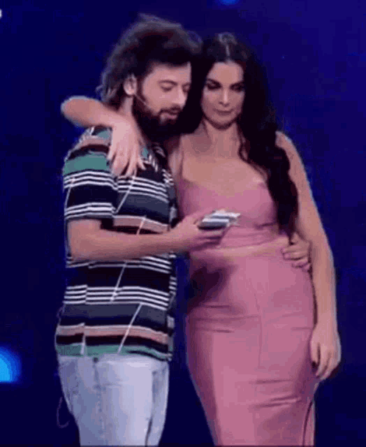 a man with a beard is hugging a woman in a pink dress while she looks at her phone .