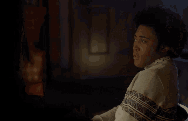 a young man in a sweater is sitting in a dark room and looking at the camera .