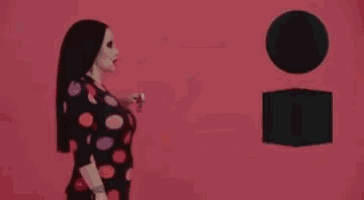 a woman in a polka dot dress is holding a glass of wine in her hand while standing in front of a pink wall .
