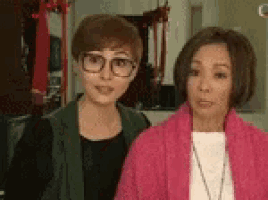 two women are standing next to each other in a room . one of the women is wearing glasses .