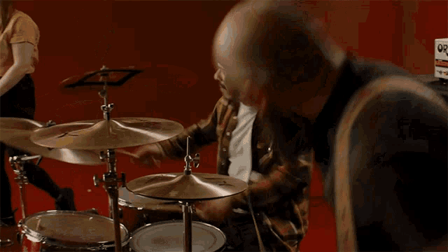 a man in a plaid shirt plays drums in front of a red wall