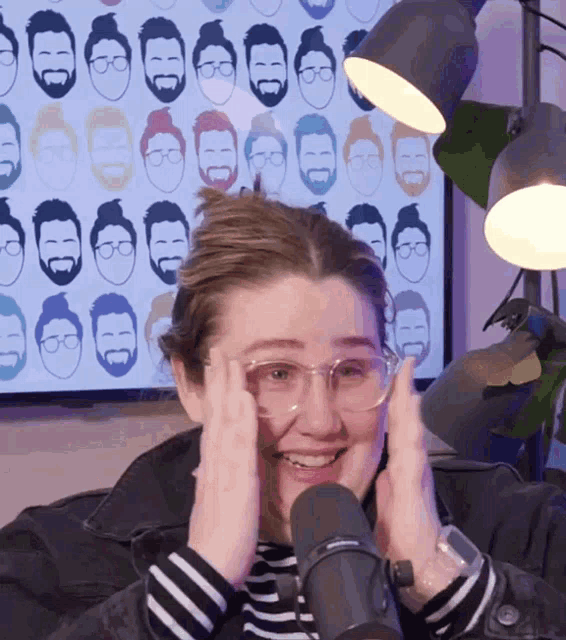 a woman wearing glasses is smiling in front of a microphone in front of a wall with bearded men 's faces