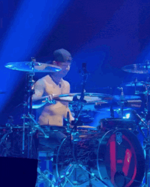 a shirtless man playing drums on a stage with a blue light behind him