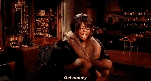 a woman in a fur coat is standing in a room talking about getting money .