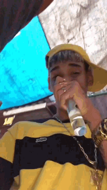 a young man wearing a yellow hat and a yellow and black striped shirt is drinking from a bottle