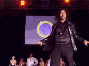 a man in a black jacket is dancing on a stage in front of a screen that says ' a ' on it