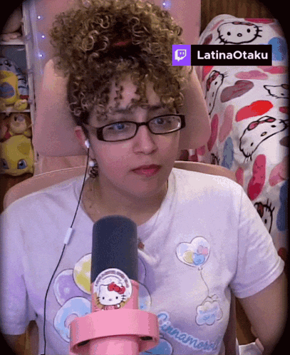a woman wearing glasses and a hello kitty shirt is sitting in front of a microphone with a twitch icon above her head