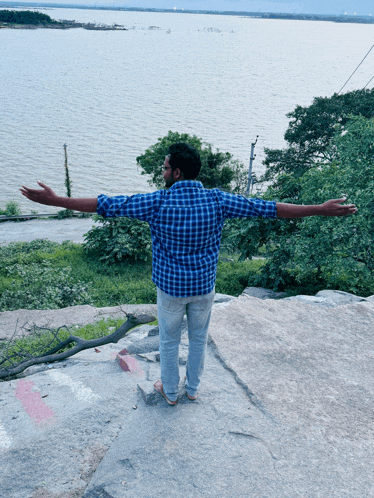 a man in a blue plaid shirt is standing on a rock with his arms outstretched in front of a body of water