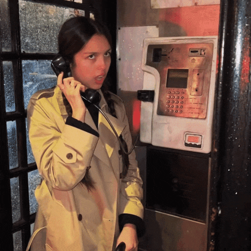 a woman in a trench coat talking on a payphone